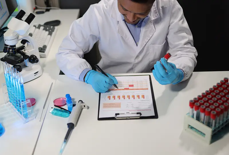 sample 4 A scientist in a lab coat writes on a clipboard while holding a test tube, surrounded by laboratory equipment such as a microscope, pipette, and racks of test tubes.